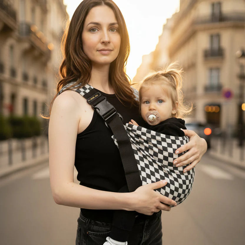 maman portant sa fille en echarpe de portage damier en ville bebe regardant la rue