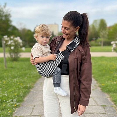 maman souriante portant son bébé en echarpe de portage pied-de-poule au parc